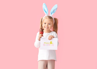 Wall Mural - Cute little girl in bunny ears with chocolate Easter egg and card on pink background