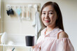 © DG PhotoStock - Good looking and smart Asian young adult woman holding a coffee mug and looking at camera with her beautiful smile in kitchen. Delightful Asian woman holding a coffee mug in kitchen portrait.