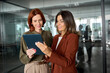 © insta_photos - Two happy busy business women talking using tab working together standing in office. Professional female colleagues employees looking at digital tablet tech discussing corporate project at work.