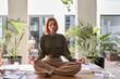 © insta_photos - Calm happy serene young professional business woman with red hair sitting on work desk meditating, doing yoga breathing exercises for no stress in modern green cozy office. Candid photo.