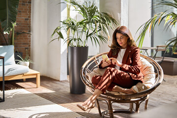 Wall Mural - Busy beautiful mature middle aged business woman using mobile cell phone looking at smartphone sitting in comfortable chair in modern office with green plants lit with sunlight. Authentic photo.