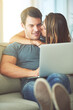© Marius Venter/peopleimages.com - Kiss, computer and woman hugging man on sofa networking on social media, website or internet. Happy, love and female person embracing husband reading online blog with laptop in living room at home.