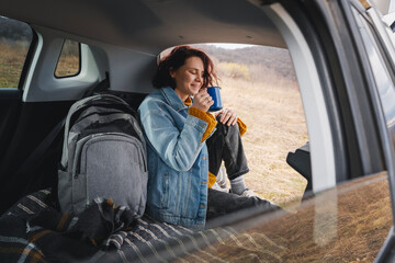 Wall Mural - A young happy woman sitting in an open car trunk drinking tea from a mug. Travel by car concept