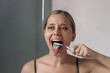 © Марина Демешко - Young caucasian blonde woman cleaning her tongue from white plaque using a toothbrush looking in the bathroom mirror. Morning routine