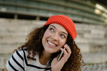 Wall Mural - pretty curly smiling woman sitting in city street in striped t-shirt and knitted red hat, using smartphone