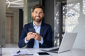 Wall Mural - Portrait of a young businessman sitting in a suit at a desk inside the office, holding a mobile phone, smiling and looking at the camera