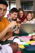 © CarlosBarquero - Vertical. Selective focus on a smiling male young university student using laptop in the cafeteria on campus with his female classmates. Academic multiracial people studying together on the faculty