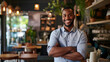 Cheerful African American male cafe owner with arms crossed, standing in his cozy coffee shop.