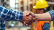 © apichat - Close-up of a firm handshake between two construction workers at a building site, representing a professional agreement.