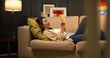 © Chanakon - Asian beautiful woman lay down on cozey couch sofa reading book in living room with warm light at night. People female stay home part time relax at home.