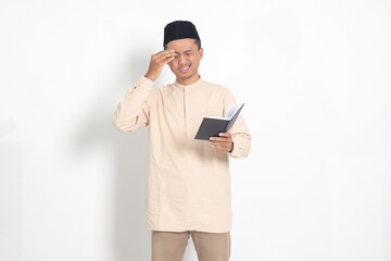Wall Mural - Portrait of confused muslim man in koko shirt with peci reading a notebook. Unhappy Asian guy holding his forehead, having headache. Isolated image on white background