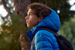 a handsome young man in profile in nature, a teenage boy in a blue puffer jacket & creme hoodie photographed as he looks out into the distance from his vantage point in amongst natural green foliage