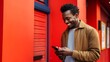 © liliyabatyrova - A smiling, happy African-American Man Uses A Smartphone, Types a message, Watch Funny Video jokes near the Red Wall on the street with a copy space. Mobile Applications, Social Networks, New Concepts