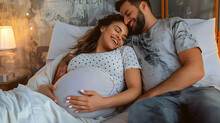 Happy Young Pregnant Wife Lying In Bed With Her Husband In The Bedroom, Man Is Lying On Her Belly