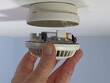 © Shaun Daley - Closeup POV of a man’s hand fitting the lower part of a smoke alarm, holding a new battery, to the upper part which is attached to a ceiling.