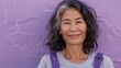 Smiling woman with gray hair wearing a gray shirt and purple suspenders standing against a purple wall with a rough texture.