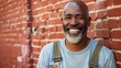 © iuricazac - Smiling man with gray beard and mustache wearing blue t-shirt and suspenders leaning against red brick wall with a warm inviting smile.