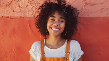 smiling woman with curly hair wearing orange suspenders over a white t-shirt against a textured oran