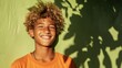A young boy with curly hair wearing an orange shirt smiling brightly against a green textured wall with shadows of leaves.