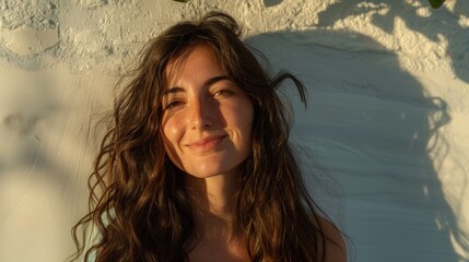 Wall Mural - A woman with long wavy hair smiling at the camera with sunlight casting a soft shadow on her face and the wall behind her.