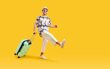 Wall Mural - Happy male tourist goes on summer vacation and travels abroad. Cheerful young man in T shirt, white trousers, hat and sunglasses with green holiday suitcase walking isolated on yellow color background