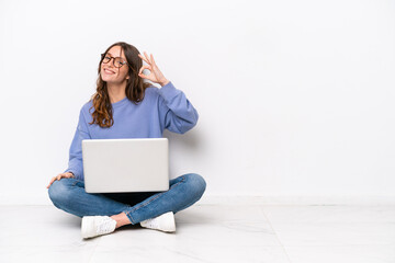 Wall Mural - Young caucasian woman with a laptop sitting on the floor isolated on white background showing ok sign with fingers