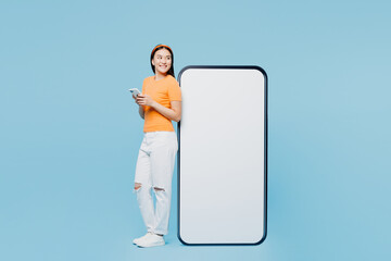 Wall Mural - Young woman of Asian ethnicity wear orange t-shirt casual clothes big huge blank screen mobile cell phone with workspace copy space use smartphone isolated on plain blue background Lifestyle concept.