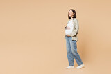 Fototapeta Panele - Full body side view young minded pregnant woman future mom in grey shirt with belly stomach stroking put hand on tummy with baby isolated on plain beige background Maternity family pregnancy concept