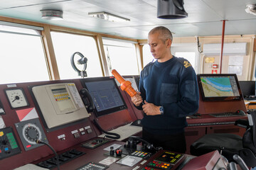 Wall Mural - Officer on watch with SART on the navigational bridge. Caucasian man in blue uniform sweater using search and rescue radar transponder on the bridge of cargo ship.