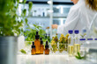 Amber bottles containing herbal extracts or essential oils on a lab table, with floral herbal samples in a clear box, and a blurred female researcher conducting extraction experiments in background