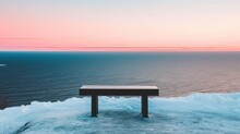 A Bench Sitting On Top Of A Pile Of Snow Next To The Ocean With A Pink Sky In The Background.