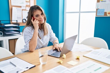 Wall Mural - Young beautiful hispanic woman business worker using laptop talking on smartphone at office