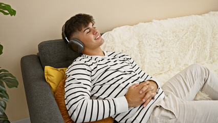 Wall Mural - Cheerful young hispanic man in headphones zone out listening to pumped music, confidently enjoying fun indoor lifestyle. he's reclining on a sofa at home, full of happiness and smiling.