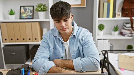 Wall Mural - Focused young hispanic man working hard, serious business worker sitting with arms crossed gesture at office desk, portrait of a successful professional boss