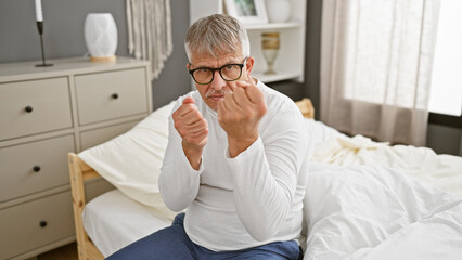 Wall Mural - Mature grey-haired man in glasses making fists while sitting on a bed in a well-lit bedroom