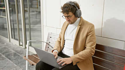 Poster - A focused young man wearing headphones works on a laptop while sitting on an urban bench outdoors.