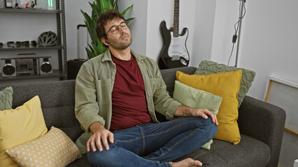 Sticker - A relaxed young man with a beard is resting on a couch in a well-decorated apartment living room with a guitar in the background.