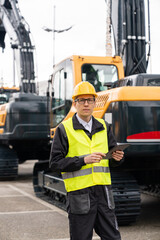 Wall Mural - Engineer in a helmet with a digital tablet stands next to construction excavators..