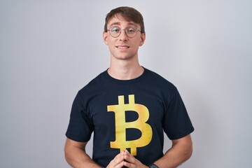 Poster - Caucasian blond man wearing bitcoin t shirt hands together and fingers crossed smiling relaxed and cheerful. success and optimistic