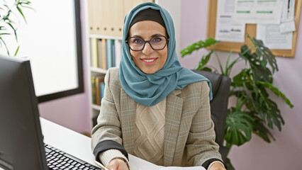 Canvas Print - Smiling middle-aged woman with glasses and hijab working in a modern office, portraying professionalism and confidence.