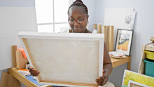 Confident African American Woman Artist Smiling, Enjoying Her Hobby, Holding Brush And Painting On Canvas While Looking At Her Artwork In Studio