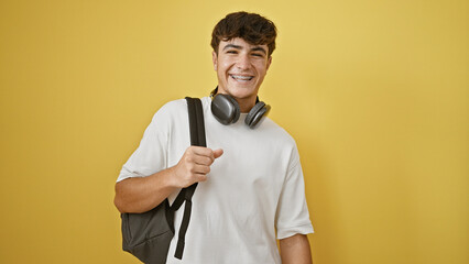 Beaming young hispanic male student with headphones and backpack, radiating sheer joy against cool yellow isolated wall