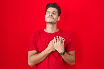 Canvas Print - Young hispanic man standing over red background smiling with hands on chest with closed eyes and grateful gesture on face. health concept.