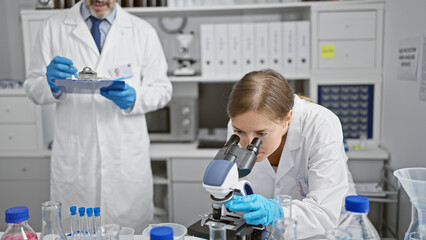 Sticker - Two serious scientists together in their lab; man and woman partners working on research, discovering through microscope, taking detailed notes on clipboard checklist