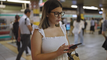 Poster - Stunning hispanic woman, glasses on, standing at underground railroad station, anticipating her subway journey, engrossed in her phone