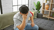 © Krakenimages.com - A young asian man in casual attire sitting dejectedly on a gray sofa indoors, with his head in hand seemingly stressed or upset