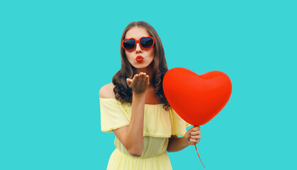 Wall Mural - Portrait of happy young woman holding red heart shaped balloon blowing kiss on blue background