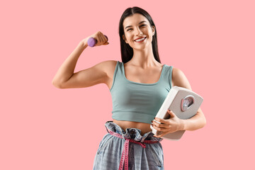 Canvas Print - Young woman in loose jeans with dumbbell and weight scales on pink background. Diet concept