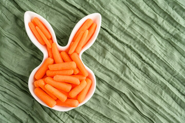 Wall Mural - Fresh raw baby carrots in an Easter Bunny shaped white ceramic dish, health and nutrition on a wrinkled pastel green gauze background
