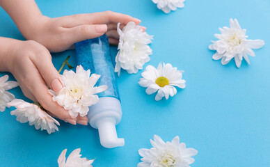 Wall Mural - Flat lay woman hands with bottle with water foam for cleansing on blue background with white flowers. Concept banner organic cosmetics for facial skin care.